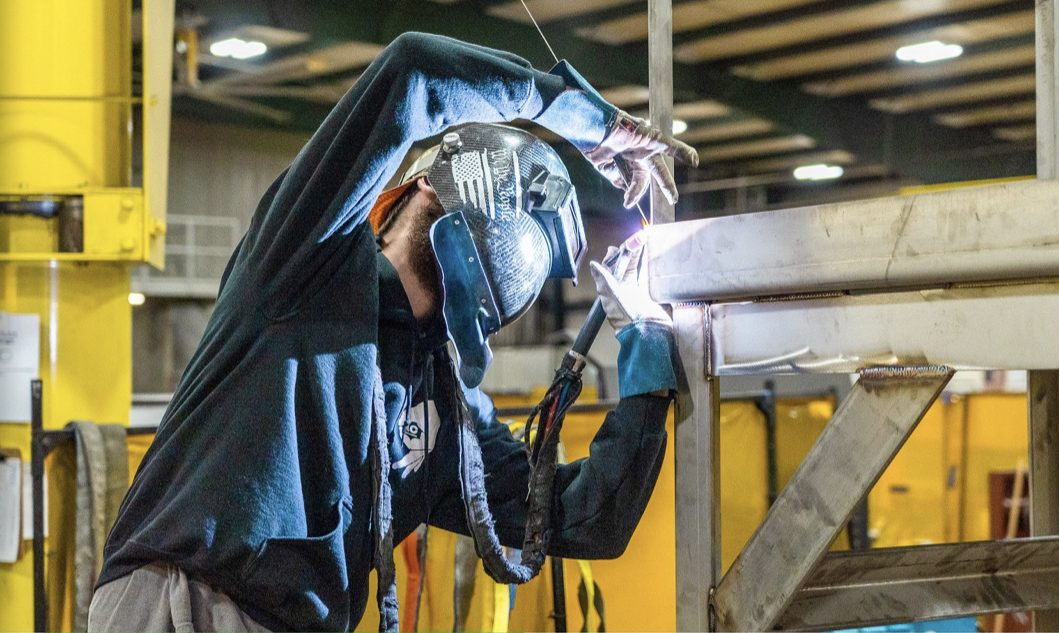 A welder in a flag-graphic hood laying a TIG bead on a stainless structural frame